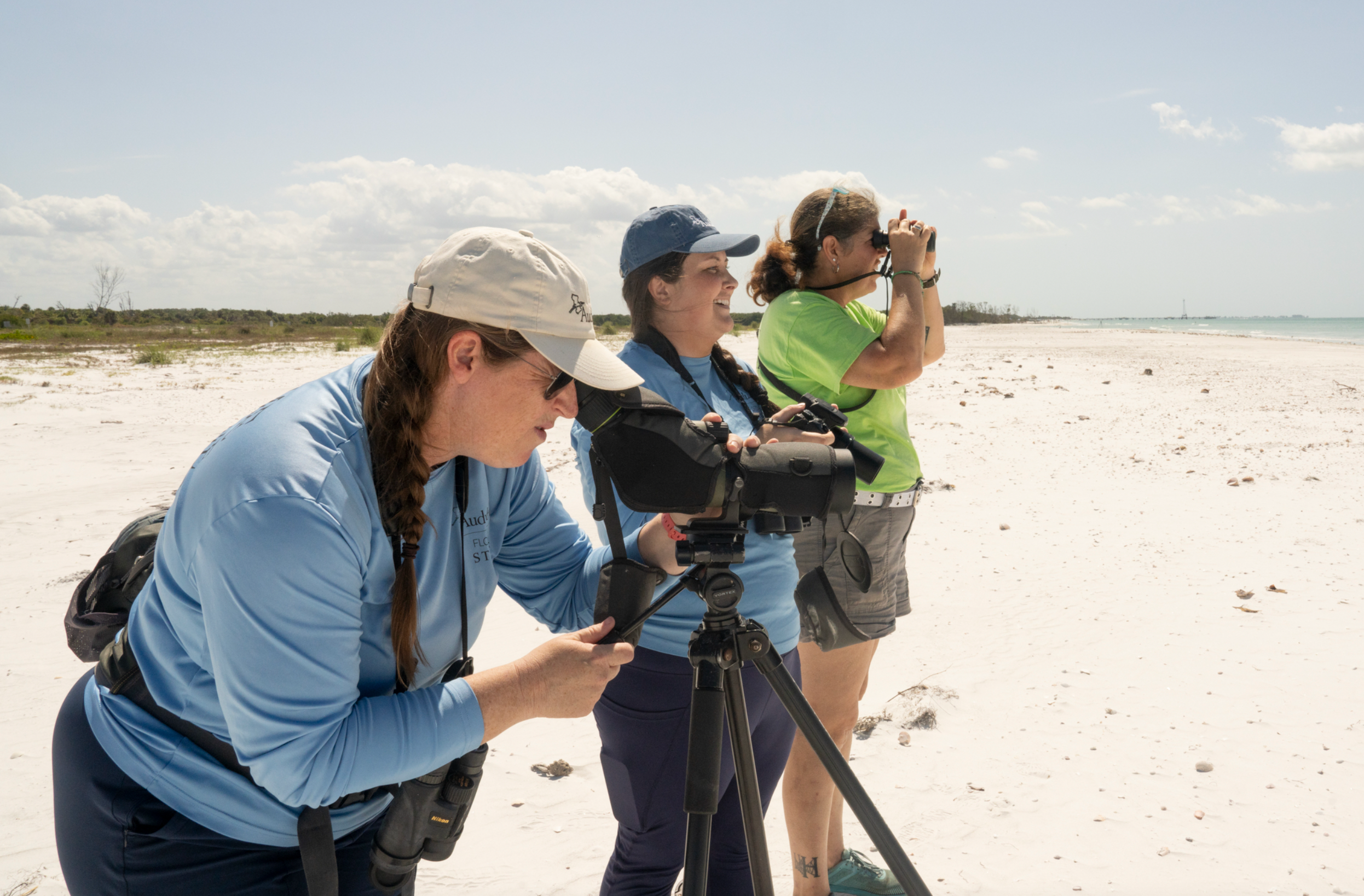 Three people looking through scopes and binoculars at the beach